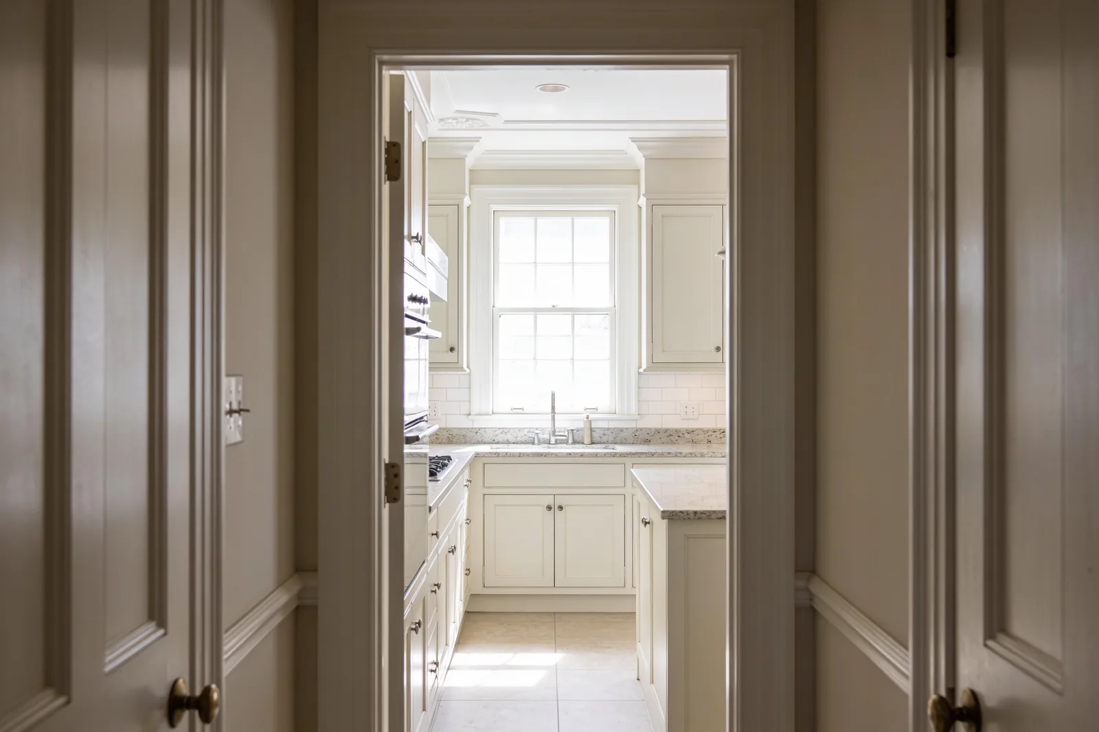 A finished kitchen seen through a hallway doorway, with the hall in shadow and daylight beyond